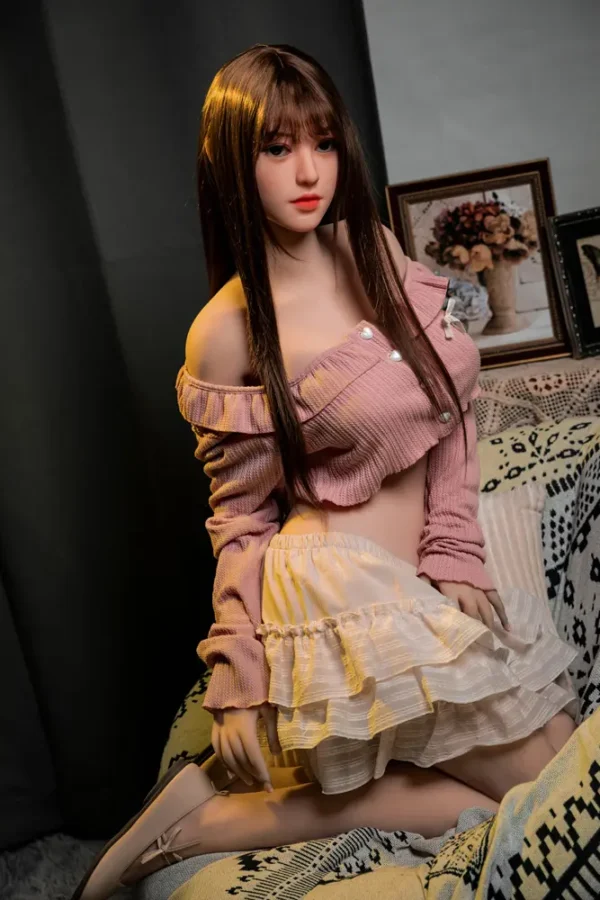 A Asian style big booty d-cup ultar realistic sex doll kneels on the sofa, wearing a pink shirt and short skirt.