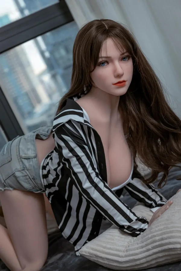 A sweet-looking sex doll kneels on a bed with her hands on a throw pillow to show off the curves of her breasts.