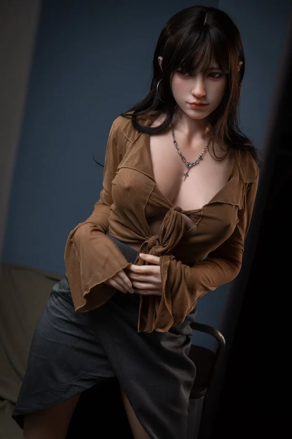 A life size d-cup real silicone head sex doll stands on the ground, wearing a brown top.