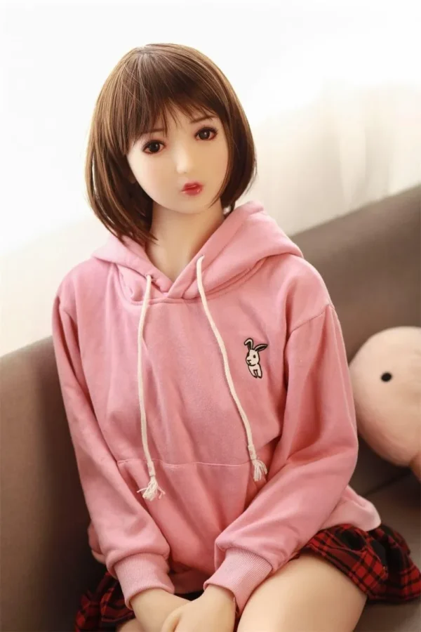 A Japanese sex doll, dressed in a pink hoodie and a red plaid skirt, sits on a sofa.