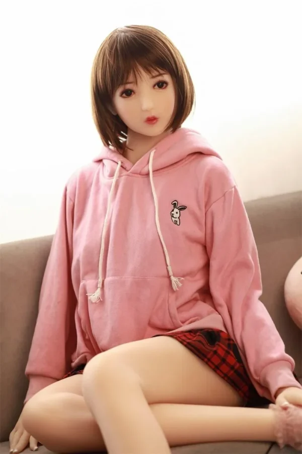 A Japanese sex doll, dressed in a pink hoodie and a red plaid skirt, sits on a sofa with her legs acrossing.