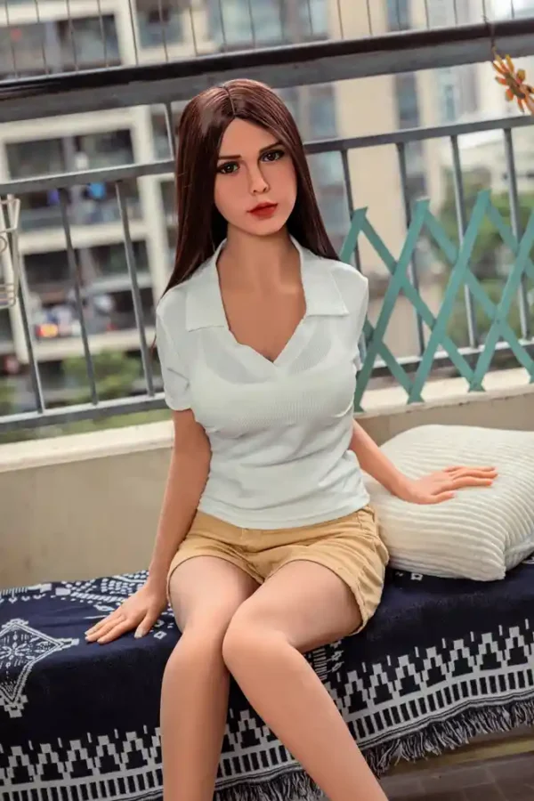 A sex doll in a light green top and yellow shorts sits on a sofa stool on the balcony with her left hand resting on a pillow and her right hand on a stool.