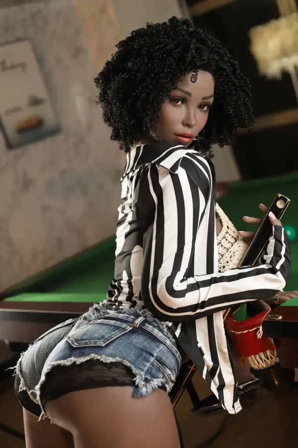 A black afro sex doll in a black and white striped coat and blue denim shorts leans over a pool table, showing off beautiful side curves.