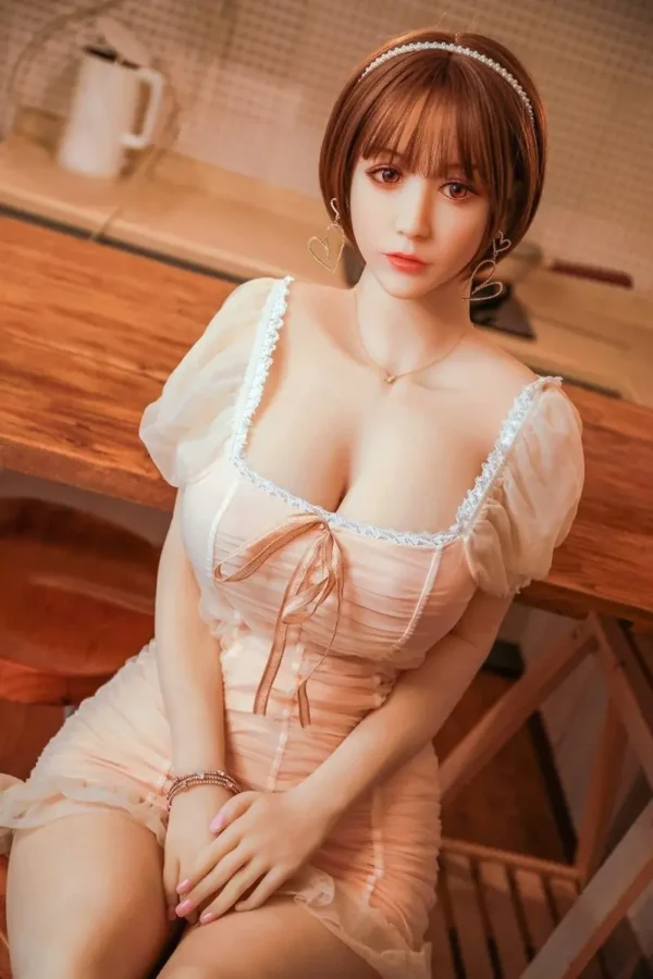 A cute young sex doll in a pink square-necked dress sits on a chair with her hands between her legs.