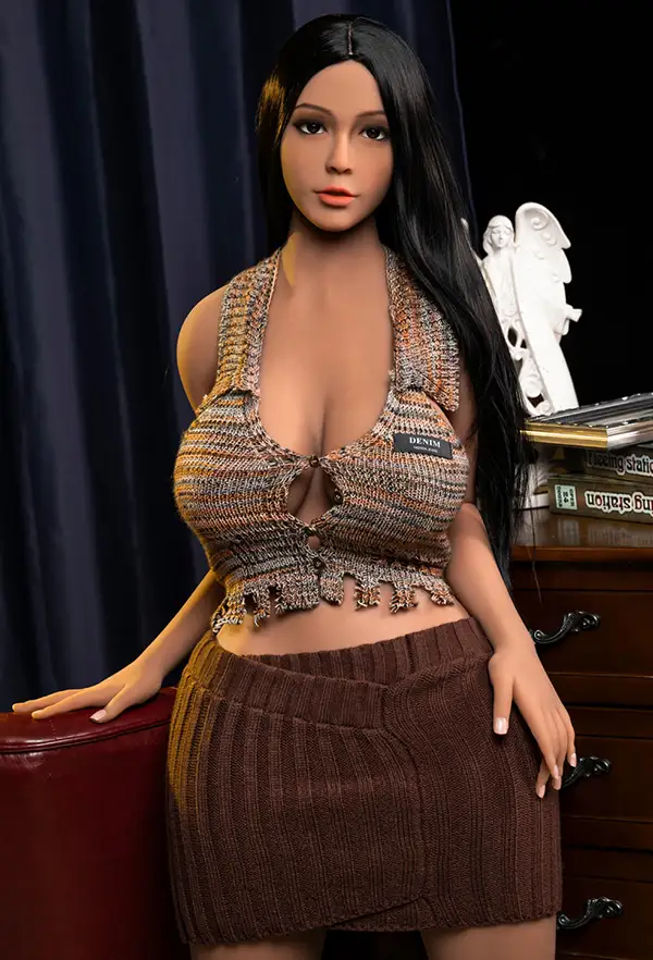 A Latina sex doll in a deep U-halter top and brown knit skirt leans against the arms of a red sofa.